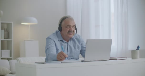 Middle-aged man is communicating by internet, using laptop and headphones, taking and making notes in copybook — Stock Video