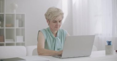 Yaşlı kadın evde dizüstü bilgisayarla çalışıyor, keskin diş ağrısı hissediyor, yanağına dokunuyor ve acıdan buruşuyor.