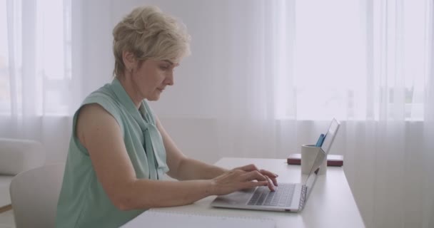 Oude vrouw is chatten op sociale netten via internet, met behulp van laptop, het typen van bericht op het toetsenbord, zitten aan tafel — Stockvideo