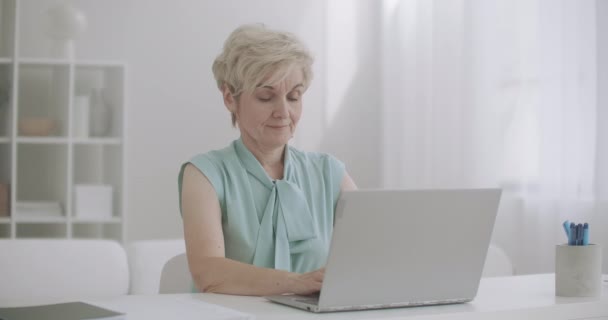 Femme âgée fatiguée étire les mains et les épaules pendant le travail avec un ordinateur portable, assis à la table et tapant des messages — Video