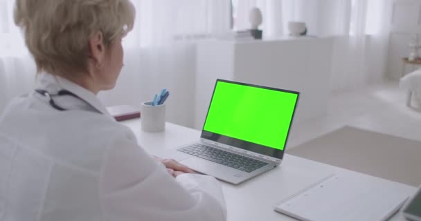 Bejaarde vrouwelijke arts werkt op afstand, online consultatie, op zoek naar groene scherm van laptop voor chroma belangrijkste technologie, telegeneeskunde — Stockvideo