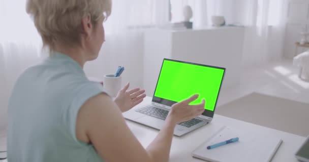 Mulher loira está se comunicando on-line por chamada de vídeo, olhando para a tela verde do laptop para a tecnologia chave chroma, gesticulando e acenando — Vídeo de Stock