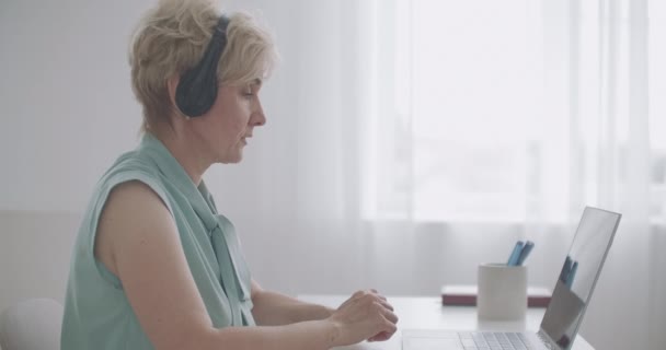 Leeftijd vrouw communiceert met collega 's of klanten via online chat op laptop, praten en luisteren via een koptelefoon — Stockvideo