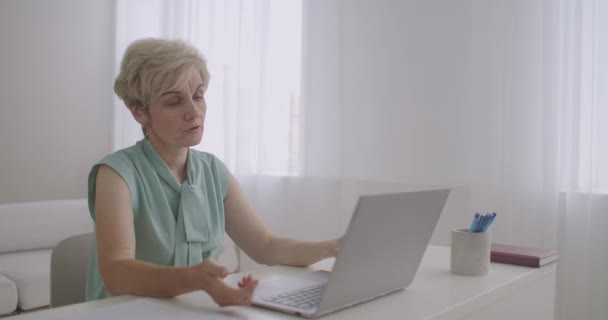 上了年纪的女教师正在网上讲课，看着笔记本电脑的网络摄像头，互联网的远距离教育，妇女的肖像 — 图库视频影像