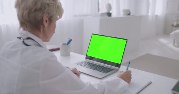 Mujer médico está viendo webinar, mirando la pantalla verde de la computadora portátil para la tecnología de croma key, escribir notas en copybook, formación avanzada — Vídeos de Stock