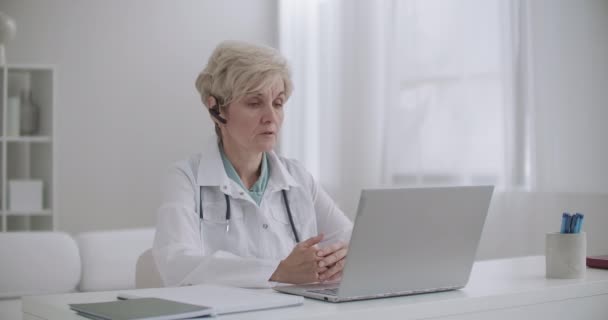 Mujer anciana pediatra está consultando en línea, sentado en la oficina del hospital y hablando con la cámara web de la computadora portátil, sesión de telemedicina — Vídeos de Stock