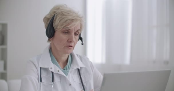 W wieku kobieta lekarz rodzinny jest konsultantem online, rozmawia z pacjentem przez czat wideo na laptopie, pracuje zdalnie — Wideo stockowe