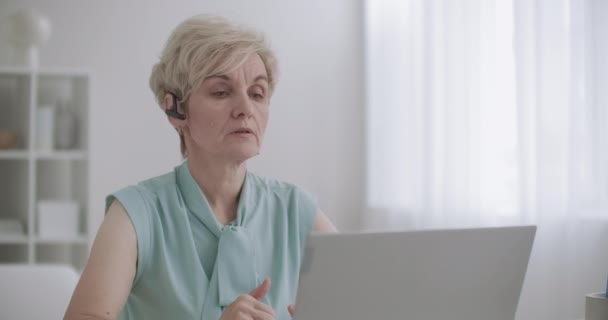 Kvinnlig specialist på call-center talar med headset och tittar på visning av bärbar dator, porträtt inomhus, arbetar på distans — Stockvideo