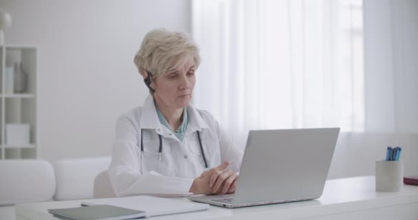 Médico chefe do hospital feminino está se comunicando com os médicos por videoconferência on-line no laptop, sentado em seu escritório — Vídeo de Stock