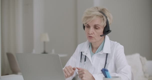 Старушка-врач читает лекции на онлайн-конференции, сидит в своем кабинете в больнице с ноутбуком — стоковое видео