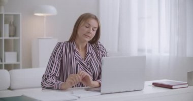 Öğrenci kadın evde dizüstü bilgisayar ve defter ile öğreniyor. Yorgun kaslarını geriyor.