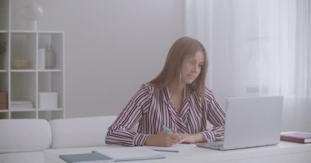 Estudiante femenina está aprendiendo en línea, ver video lección en el ordenador portátil y escribir en el libro de ejercicios, e-educación para los jóvenes — Vídeo de stock