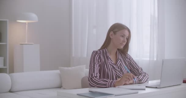 Joven bonita estudiante mujer está aprendiendo en línea en casa, sentado en la mesa, viendo webinar en el ordenador portátil y escribir notas — Vídeo de stock