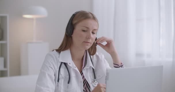 Jonge vrouwelijke arts communiceert met de patiënt online via laptop, met behulp van videocall en koptelefoon met mic — Stockvideo