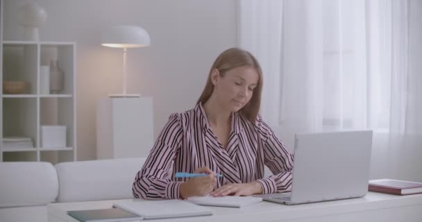 在家里上网学习的时候，年轻的女人正在看笔记本电脑的展示，在抄本上写字，在遥远的教育 — 图库视频影像
