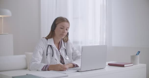 Terapeuta joven está consultando en línea, pidiendo paciente y tomando notas en copybook, trabajando remotamente — Vídeo de stock