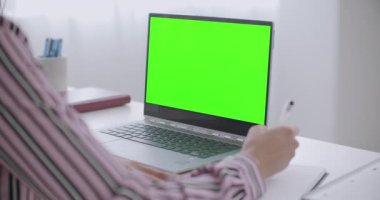 Genç kadın öğreniyor, defterde yazıyor ve krom anahtar teknolojisi için dizüstü bilgisayarda yeşil ekrana bakıyor.