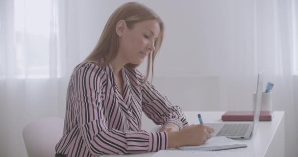 Estudiante mujer está aprendiendo en línea, viendo webinar en el ordenador portátil y tomando notas en copybook — Vídeo de stock