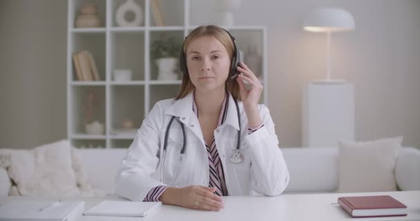 Junge Ärztin hört Patientin oder Kollegin per Kopfhörer zu, nutzt Videoanrufe, nickt in die Kamera — Stockvideo