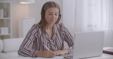 Kulaklıklı ve mikrofonlu genç bir kadın laptop 'daki video konferansında sohbet ediyor, evden uzaktan çalışıyor, internet teknolojisi