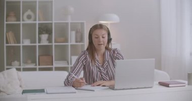 Genç bir kadın uzaktan çalışıyor, internetteki görüşmeleri bilgisayardaki görüntülerle yürütüyor, mikrofonlu kulaklıklar kullanıyor.