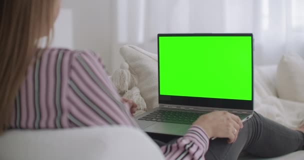 Jovem está sentado no sofá e segurando laptop com tela verde para a tecnologia chave chroma, conversando por chamada de vídeo — Vídeo de Stock