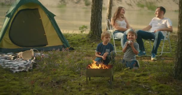 Sebuah keluarga di alam, orang tua menonton sebagai dua anak laki-laki di api panggang marshmallow pada tongkat di latar belakang tenda. Tenda tenda sebagai keluarga — Stok Video