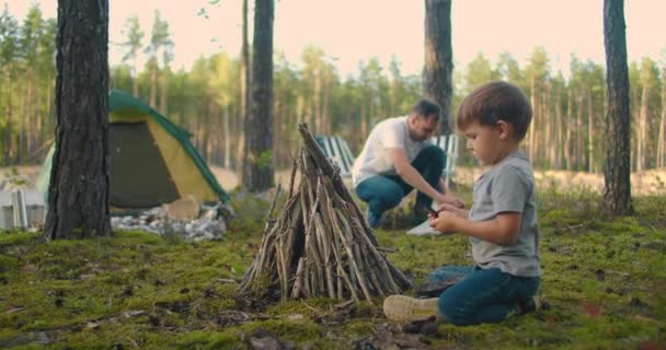 Een jongen van 3-4 jaar helpt om stokken in een kampvuur te steken tijdens een reis naar het bos als gezin in de natuur. Familie vakantie in het bos met tenten — Stockvideo