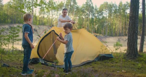 Den unge mannen och två unga pojkar satte tillsammans upp ett tältläger för natten under kampanjen. Faderskap och en lycklig barndom. Far och söner slår upp ett tält tillsammans i skogen — Stockvideo