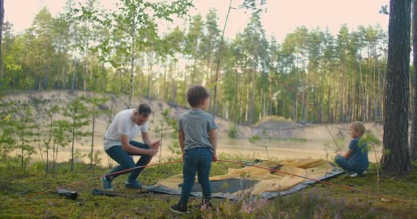 Der junge Mann und zwei junge Burschen bauten während der Aktion gemeinsam ein Zeltlager für die Nacht auf. Vaterschaft und eine glückliche Kindheit. Vater und Söhne bauen gemeinsam ein Zelt im Wald auf — Stockvideo