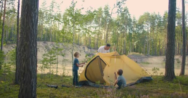 De jongeman en twee jonge jongens zetten samen een tentenkamp op voor de nacht tijdens de campagne. Vaderschap en een gelukkige jeugd. Vader en zonen zetten samen een tent op in het bos. — Stockvideo