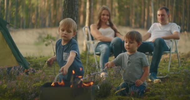 Due ragazzi 3-6 anni insieme friggono in fuoco marshmallows su bastoni sullo sfondo di genitori. Escursione in famiglia nel bosco con tenda. Famiglia in natura in escursione — Video Stock