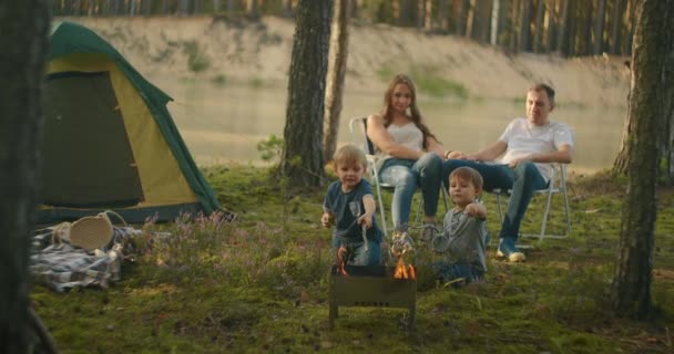 Sebuah keluarga di alam, orang tua menonton sebagai dua anak laki-laki di api panggang marshmallow pada tongkat di latar belakang tenda. Tenda tenda sebagai keluarga — Stok Video
