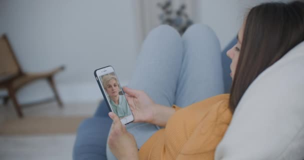 自宅の携帯電話の画面上でカンファレンスチャットオンラインアプリケーションを使用して、古い親の母親や成熟した友人を呼び出す若い女性の娘のビデオの肩のビュー以上。ファミリービデオコンセプト — ストック動画