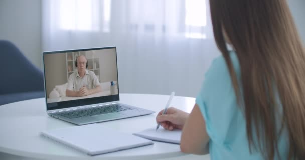 Studente della scuola della ragazza a distanza che impara agitando la mano che studia con l'insegnante online sullo schermo del computer portatile. Elearning zoom videochiamata, lezione di videoconferenza con tutor. Vista sulla spalla. — Video Stock