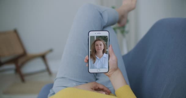 Σγουρή νεαρή γυναίκα που κάνει βιντεοκλήση με φίλους χρησιμοποιώντας κάμερα κινητού τηλεφώνου ενώ είναι ξαπλωμένη στο κρεβάτι. Ελκυστική χαρούμενη γυναίκα κάθεται στον καναπέ και έχει ένα βίντεο κλήση με φίλες — Αρχείο Βίντεο