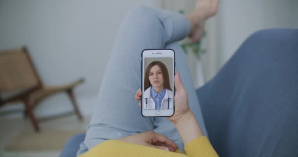 Жінка лежить на дивані, розмовляючи з лікарем на відео за допомогою смартфона. Віддалена медична консультація з лікарем через мобільний телефон — стокове відео