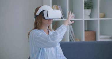 Sanal gerçeklik kulaklığı takan neşeli genç bir kadın evde yatağında oturmuş 360 VR video filmi izliyor. Sanal gerçeklikteki genç kadın portresi evdeki koltukta oturuyor.