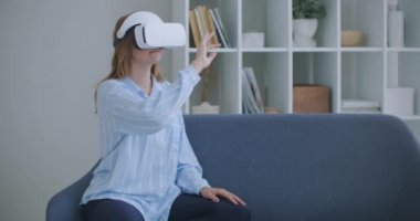 Sanal gerçeklik kulaklığı takan neşeli genç bir kadın evde yatağında oturmuş 360 VR video filmi izliyor. Sanal gerçeklikteki genç kadın portresi evdeki koltukta oturuyor..