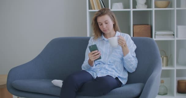 Menina adolescente ruiva atraente sentado do sofá em casa usando telefone e sorrindo. Sinto-me feliz. Comunicação feminino olhando mensagem no telefone celular ou smartphone. — Vídeo de Stock