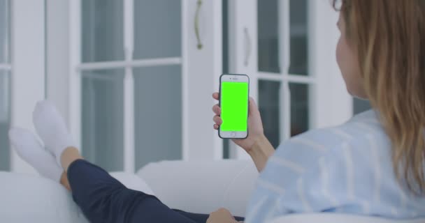 Foto sobre el hombro de una mujer sosteniendo un teléfono móvil o teléfono inteligente con una pantalla verde vertical en la mano en su casa tumbada en el sofá. Chica toque en la pantalla y ver contenido. — Vídeos de Stock