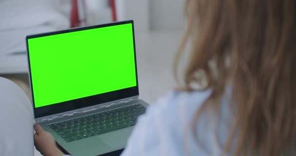 Mladá žena doma pracuje na notebooku se zelenou maketou. Sedí na gauči v jeho útulném obýváku. Přes rameno Kamera Shot