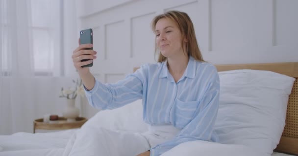 Liikenainen, jolla on videopuhelu hotellissa. Onnellinen tyttö makaa sängyllä, puhuu ystävien kanssa videopuhelun kautta. — kuvapankkivideo