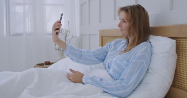 Bizneswoman ma rozmowę wideo w hotelu. Szczęśliwa dziewczyna leży na łóżku, rozmawia z przyjaciółmi przez telefon wideo. — Wideo stockowe