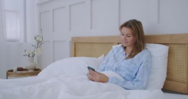 Çekici bir genç kız yatak odasında otururken arkadaşlarıyla sohbet ediyor. Genç bir kadın yatakta uzanıyor ve elinde akıllı telefon tutuyor.