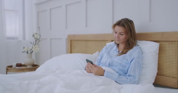 Atractiva joven hermosa chica charlando con sus amigos, mientras está sentada en su dormitorio. Mujer joven acostada en la cama y sosteniendo el teléfono inteligente — Vídeos de Stock