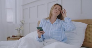 Çekici bir genç kız yatak odasında otururken arkadaşlarıyla sohbet ediyor. Genç bir kadın yatakta uzanıyor ve elinde akıllı telefon tutuyor.