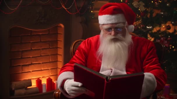 Hombre mayor barbudo en traje de cláusula santa hojeando páginas de libro con cubierta roja en habitación temáticamente decorada - concepto de espíritu navideño de cerca. — Vídeos de Stock