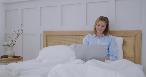 Fiatal Y-generációs lány ül az ágyon reggel, Laptop számítógépet használ. Kaukázusi nő otthon városi dolgozik online laptop hálószoba ágy — Stock videók