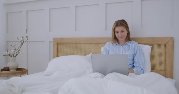 Mulher feliz com laptop deitado na cama e sorrindo. menina em uma camisa de homem trabalhando em um laptop no início da manhã — Vídeo de Stock